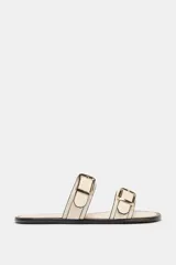 Sandalia plana de color beige con dos tiras ajustables mediante hebillas metálicas doradas. Presenta un diseño de punta redonda y suela plana con borde en contraste.