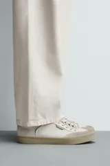 Zapatilla deportiva de estilo retro, color blanco con detalles en gris y negro. Presenta una suela de color beige en contraste y cordones blancos.