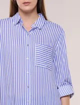 Camisa de corte clásico y silueta relajada con diseño de rayas verticales en blanco y negro. Presenta cuello camisero tradicional, cartera frontal con botones, un bolsillo aplicado en el pecho con rayas horizontales y mangas largas con sistema para arremangar.