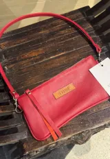 Minicartera de cuero color rojo de la marca Soriame, con diseño rectangular, cierre superior y tira larga ajustable. Cuenta con una etiqueta frontal de cuero con el logo grabado y tiras colgantes decorativas.