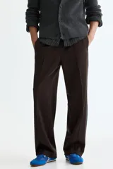 Pantalón deportivo marrón de punto con leve brillo, corte relajado y piernas rectas con pliegues cosidos. Cuenta con franjas laterales oscuras y ribetes blancos contrastantes, cintura elástica y bolsillos laterales.