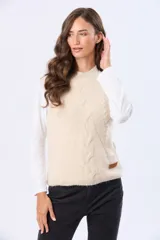 Chaleco tejido de punto grueso en color beige, con cuello redondo y diseño estructurado con detalles de trenzas verticales. Presenta un pequeño aplique de cuero sintético en la parte inferior.