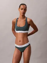 Bralette deportivo sin relleno ni forro, con diseño de espalda cruzada y banda elástica inferior con el logo de la marca. Presenta un estampado geométrico con el monograma de Calvin Klein en tonos verde oscuro y negro.