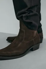 Bota estilo cowboy de caña media, confeccionada en piel con acabado de gamuza en color marrón oscuro. Presenta horma en punta, cierre lateral y suela con tacón cuadrado bajo.