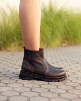 Bota de caña alta confeccionada en cuero color marrón caoba con acabado desgastado. Presenta una suela track robusta, correas con hebillas en la parte superior de la caña y un arnés decorativo con herrajes metálicos en el tobillo.