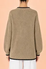 Cardigan de punto en color beige con textura tejida, cuello redondo y cierre frontal de botones. Presenta un detalle de borde festoneado en color negro en el cuello, la tapeta de botones, el ruedo y los puños.