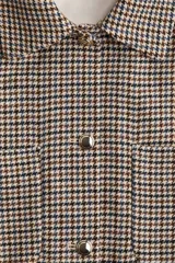 Chaqueta de tejido grueso con estampado pata de gallo en tonos beige, marrón y negro. Presenta cuello camisero, cierre frontal con botones metálicos plateados, y dos bolsillos de parche con solapa y botón en el pecho. El corte es holgado y ligeramente corto, con mangas largas.