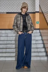 Chaqueta de estilo camisa a cuadros en tonos beige y marrón, con cierre frontal, cuello camisero y capucha integrada. Presenta un diseño holgado y mangas largas.