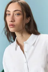 Camisa blanca de corte oversize con cuello camisero y mangas largas. Presenta un bordado en la espalda con el diseño de un escudo nacional estilizado en color azul y la frase 'El país de los mates y las siestas'.