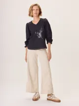 Remera de manga larga color negro con cuello en V y puños fruncidos. Presenta un estampado sutil en tonos grises y blancos en el centro del pecho, simulando un ramillete de flores secas.