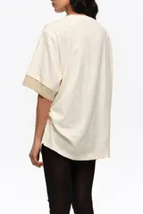 Remera larga de color marfil con detalle de abertura y lazos en el hombro, estampa frontal de zapatillas de ballet con lazos rojos y mangas cortas con puños en contraste rojo y beige. Cuenta con cordón ajustable lateral.