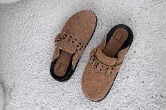 Mules de gamuza color tostado con diseño de tira ajustable con hebilla metálica y detalles de ojalillos decorativos. Posee plantilla ergonómica con logo grabado y suela plana de goma negra.