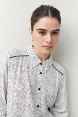 Camisa de manga larga confeccionada en 100% viscosa, color blanco con estampado de rayas finas negras. Presenta cuello camisero, botones frontales negros y detalles de ribete negro en los hombros.
