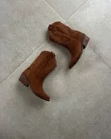 Bota corta de gamuza color marrón topo, estilo western, con bordados decorativos en hilo blanco en caña y empeine. Posee taco cuadrado de madera y punta ligeramente afilada.