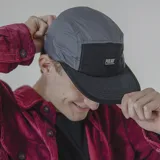 Gorro estilo five-panel de nylon, bicolor, con paneles superiores grises y panel frontal y visera negros. Presenta un parche rectangular negro con el logo 'PULAU' en blanco.