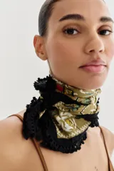 Pañuelo cuadrado de algodón con estampado floral y geométrico sobre fondo verde oliva. Presenta un borde negro con flecos decorativos en todo su contorno.