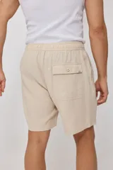 Bermuda de corte recto en color beige, confeccionada en una mezcla de algodón, lino y poliéster. Presenta cintura elástica con cordón ajustable, bolsillos laterales y una pequeña etiqueta decorativa en el ruedo.