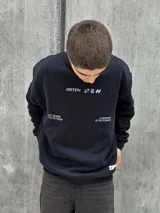 Buzo negro de corte holgado con cuello redondo y puños acanalados. Estampado frontal con la palabra "OSTEN" y la frase "NOT WHERE YOU'RE FROM A MINDSET OF BECOMING" en color blanco.