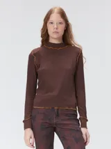 Polera de manga larga confeccionada en tela rib color marrón, con cuello alto y hombreras. Presenta detalles de costuras con pespuntes en hilo de color contrastante en el cuello, hombros, puños y ruedo.