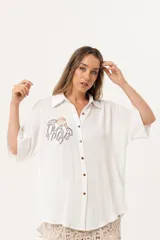 Camisa blanca de silueta oversize, con cuello camisero, manga 3/4 y corte más largo en la espalda. Presenta bordados de coral y caracol en el frente, y botones a tono.