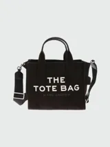 Bolso tipo tote de lona color negro con estampado frontal en blanco que dice 'THE TOTE BAG' y 'MARC JACOBS'. Cuenta con doble asa superior, correa ajustable y extraíble para llevar al hombro y cierre superior con cremallera.