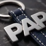 Llavero de acero con diseño de letras que forman la palabra 'PAPÁ', unido a una correa de cuero sintético azul y una argolla circular metálica.
