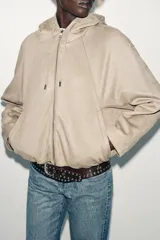 Chaqueta bomber de efecto ante en color marrón, con capucha ajustable mediante cordones, cierre frontal con cremallera, bolsillos laterales y bajo con elástico interior.