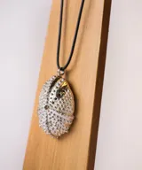 Collar con dije en forma de erizo de mar con textura granulada, suspendido de un cordón negro. El dije presenta un diseño orgánico con pequeños orificios y una superficie detallada.