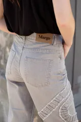 Jean gris lavado, estilo mom fit, con aplique fruncido en forma de espiral en la pierna izquierda, tiro alto, bolsillos delanteros y traseros funcionales, y cierre frontal con botón y cremallera.