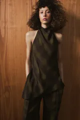 Top de cuello halter con espalda descubierta, confeccionado en tejido de viscosa con estampado de cuadros en tonos verde oliva y negro. Presenta un diseño asimétrico en el bajo y cierre con botones en la parte posterior del cuello.