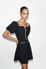 Vestido corto de tejido bouclé en color negro, con escote recto y mangas cortas abullonadas. Presenta detalles de vivos en color blanco a contraste en el escote, el centro del delantero y en los falsos bolsillos frontales.
