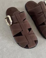 Zueco tipo sandalia de gamuza color marrón, con diseño de tiras entrecruzadas y hebilla metálica dorada lateral.