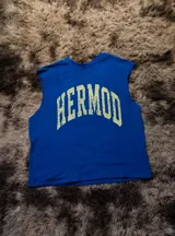 Musculosa de corte boxy, color azul vibrante, con cuello redondo y sisas amplias. Estampado frontal arqueado con la palabra "HERMOD" en tipografía universitaria desgastada color amarillo claro.