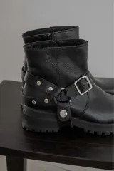 Bota corta de cuero negro con punta cuadrada, suela track y detalle de estribo desmontable con hebilla metálica.