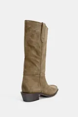 Bota de caña alta confeccionada en serraje, con diseño pull-on de calce fácil. Presenta detalle de doble tirador superior, pespuntes decorativos en el cuerpo, tacón bajo en bloque y punta cuadrada.