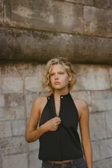 Top chaleco negro satinado con cuello halter, espalda descubierta y cierre frontal con cremallera y botón a presión ocultos por solapa.