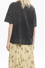 Camiseta de algodón color blanco hueso, de corte boxy y holgado, con escote redondo y mangas cortas. Presenta un pequeño logo en relieve de color rojo en el área del pecho.