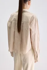 Camisa de calce relajado confeccionada en algodón, con diseño de rayas verticales en tonos beige y blanco. Presenta cuello clásico, cierre frontal con botones, puños abotonados y largo ligeramente cropped.