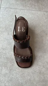Sandalias de cuero marrón con punta cuadrada, tiras texturizadas con detalles de ojales metálicos y cordones entrelazados, y tira ajustable con velcro en el talón.