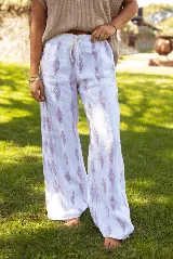 Pantalón de lino de corte amplio y tiro medio, con cintura elástica y cordón ajustable. Presenta un estampado lineal con motivos decorativos en tonos violetas sobre fondo blanco.