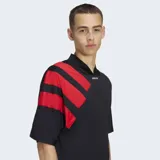 Remera Adidas de estilo futbolístico inspirada en los años 90, color negro con cuello en pico y tres bandas diagonales rojas sobre el hombro derecho. Presenta un corte holgado y logo de la marca en el centro del cuello.