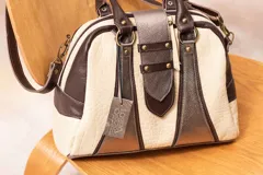 Cartera estructurada con diseño tipo bowling, combinando paneles de cuero color crema con textura cocodrilo, cuero liso marrón chocolate y paneles metálicos color peltre. Posee asas de mano y correa ajustable desmontable.