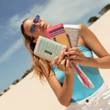 Billetera mediana con cierre perimetral, confeccionada en cuero sintético rosa y detalle central de tejido tipo telar con rayas verticales en amarillo, rosa y negro. Incluye correa de mano desmontable y placa metálica con logo de la marca.