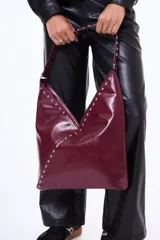 Bolso tipo hobo de cuero sintético negro con acabado brillante, decorado con tachuelas plateadas a lo largo de la correa y en el borde frontal. Presenta un diseño asimétrico en la parte superior.