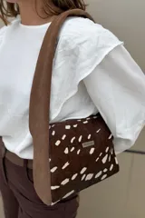 Cartera de mano tipo bandolera confeccionada en cuero con pelo vacuno estampado con manchas blancas sobre fondo marrón. Presenta una correa ancha de gamuza en color habano y una placa metálica con el logo de la marca en el frente.