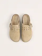 Zueco de gamuza color beige, con diseño de punta cerrada y talón descubierto. Presenta una tira ajustable sobre el empeine con hebilla metálica y ojales decorativos. Cuenta con una suela de corcho y base de goma antideslizante.