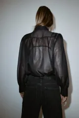 Camisa de manga larga con cuello solapa, confeccionada en tejido semitransparente con hilo metalizado. Presenta volantes frontales decorados con abalorios y cierre frontal con botones ocultos.