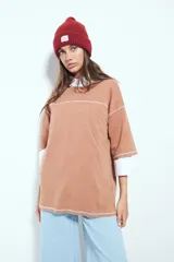 Remera de manga corta color marrón con efecto desgastado, corte oversize y costuras a la vista en color blanco. Presenta cuello redondo con cuello polo blanco superpuesto y mangas largas a rayas horizontales marrones y blancas por debajo.