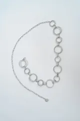 Cinturón tipo cadena de color plateado, compuesto por una serie de eslabones circulares de distintos tamaños unidos entre sí, con cierre de mosquetón y cadena de ajuste final.