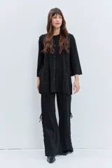 Cardigan negro de punto con acabado brillante, cuello redondo, cierre frontal de botones y detalle de cordones laterales entrelazados. Presenta mangas tres cuartos con diseño acampanado.